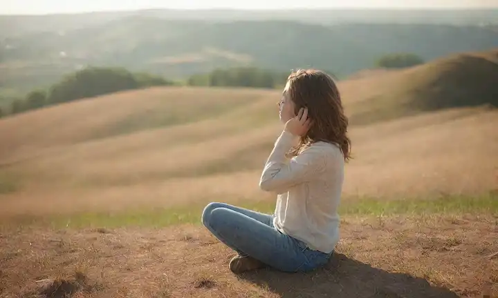 Una persona sentada en una colina, las manos detrás de la cabeza, con una expresión tranquila y pacífica; una brisa suave frotando su cabello; paisaje circundante con colores suaves y relajantes; luz del sol caliente echando sombras largas