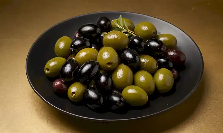 Una placa de aceitunas surtidas, verde vibrante y negro, dispuesta artísticamente en forma curvada, con un fondo sutil gradiente, ramas de olivo brotando, luz dorada que vierte desde arriba, texturas naturales, ambiente rústico