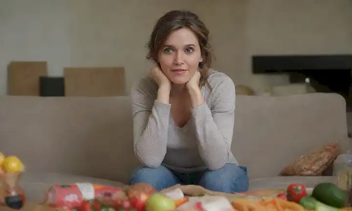 Una mujer con un peinado desordenado se sienta en un sofá, mirando estresada e infeliz, rodeada de envoltorios de aperitivos insalubres y botellas vacías, transmitiendo malos hábitos nutricionales, mientras que en el fondo una persona con pelo sano y brillante se ve sonriente, sentado cerca de fruta fresca y verduras