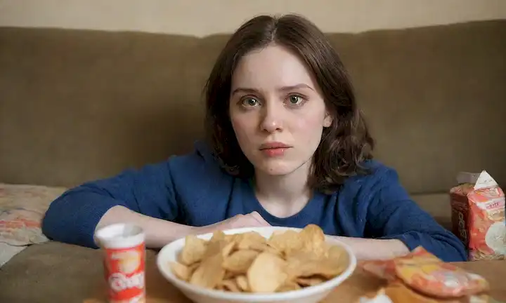 Una persona con cabello aburrido, sin vida, piel pálida y ojos hundidos, sentado en un sofá desgastado, rodeado de envolturas insalubres y botellas vacías, con una sonrisa débil insinuando la desnutrición; una inyección de un plato lleno de comida chatarra, una lata de soda y una bolsa de patatas fritas en el fondo