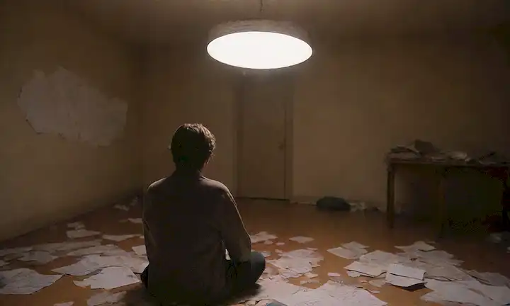 Una persona sentada sola en un cuarto iluminado con una burbuja de pensamiento sobre su cabeza, rodeada de papeles dispersos y objetos rotos, transmitiendo sentimientos de incertidumbre y confusión