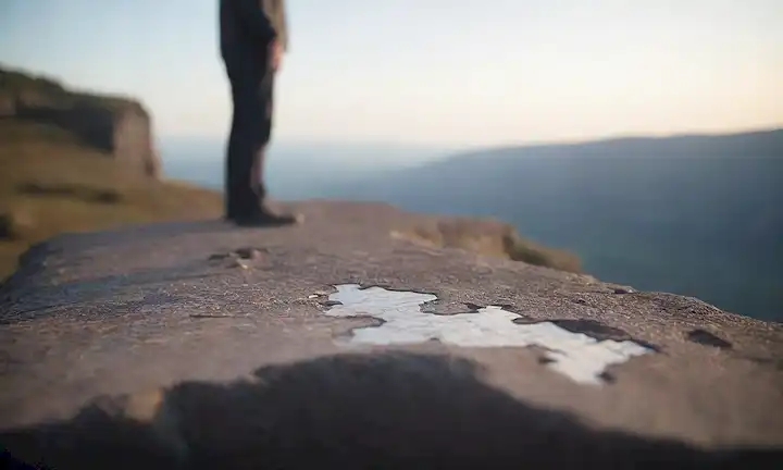 Una persona de pie en un acantilado de roca con vistas a un vasto paisaje con movimiento borroso en el fondo, transmitiendo un sentido de incertidumbre e inestabilidad; una imagen débil de una pieza de rompecabezas que encaja en el lugar; una sombra sutil de una figura que se arrastra justo fuera de la vista; una pista de caos en el entorno circundante