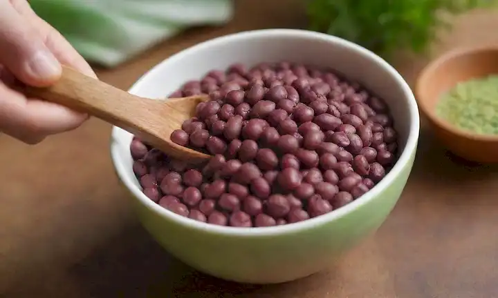 Un fondo verde vibrante, un tazón lleno de frijoles azuki, un espolvor de semillas de sésamo, unas cuantas rebanadas de fruta fresca, una mano que sostiene una cuchara, luz natural cálida, un ambiente acogedor