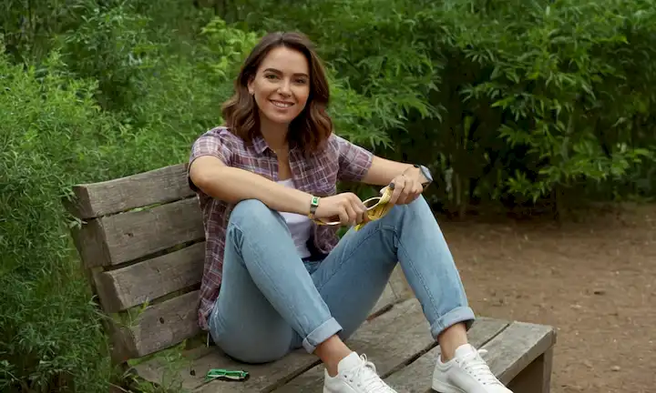 Modelo 3D de una persona en vaqueros con ajuste relajado, sosteniendo una cinta de medición y sonriendo, rodeado de vegetación, con zapatillas y camisa casual, sentado en una roca o un banco, mirando directamente al espectador