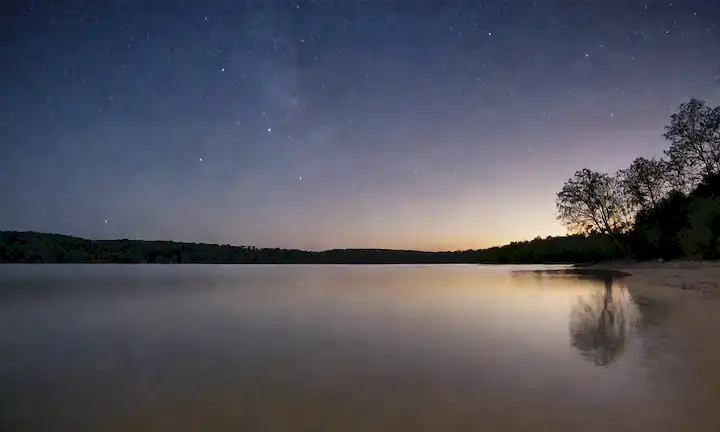 Un paisaje sereno con un tranquilo cielo nocturno, un lago tranquilo o un océano, y una playa de arena, con suaves olas y algunas estrellas, con una silueta de árboles en el fondo, transmitiendo una sensación de tranquilidad, relajación y somnolencia, todo enmarcado por un enfoque suave