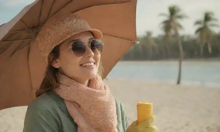 una persona que lleva un sombrero y gafas de sol en una playa con palmeras, aplicando protector solar a su nariz, sosteniendo un paraguas contra el viento frío, sonriendo con expresión facial relajada, usando ropa protectora como guantes y bufanda, sentado cerca de un cuerpo de agua