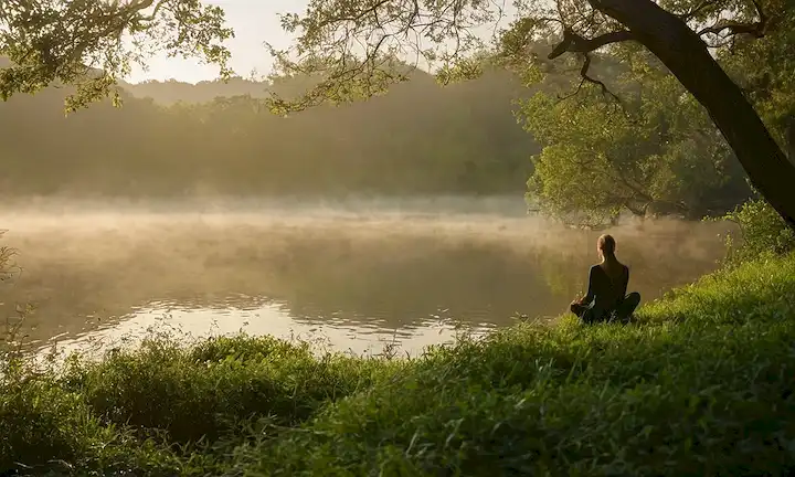 Un entorno natural sereno con unos pocos árboles de peluche, un cuerpo tranquilo de agua y una bruma sutil, con una persona en una pose meditativa en medio de una vegetación exuberante, con una luz suave y cálida iluminando la escena, transmitiendo relajación, serenidad y equilibrio