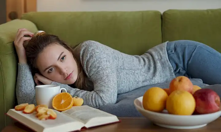 Una jovencita acostada en un sofá, con aspecto cansado y pálido, con una bebida caliente cerca, rodeada de frutas y aperitivos saludables, quizás con un libro o una revista para distraerla de la incomodidad