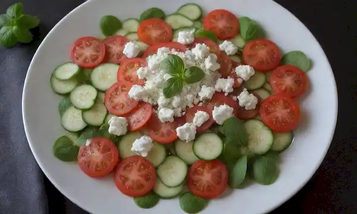 Un refrescante plato de verano con carpaccio de calabacín finamente rebanada, tomates maduros a la mitad, hojas de albahaca frescas dispersas, queso defeta desmoronado espolvoreado, flores comestibles adornadas, y un ligero goteo de aceite de oliva brillando
