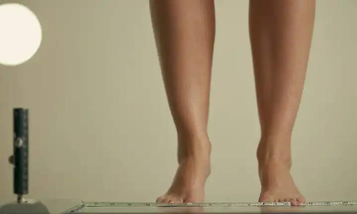 piernas con inflamación visible, gotas de agua en la piel, mujer de pie cerca de una escala o cinta de medición, fondo borroso, colores neutros, iluminación sutil, sombras mínimas