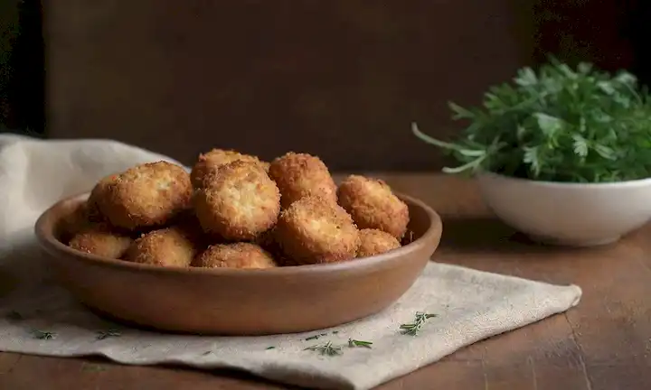 Una cálida y natural escena de luz con un tazón de croquetas fritas crujientes dispuestas artísticamente en una mesa de madera rústica, rodeada de hierbas frescas como perejil y romero, con un toque de queso dorado en el fondo, y un simple lienzo de lino como la base
