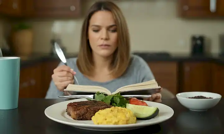 Un plato de comida con una parte de ella removida, una persona que sostiene un tenedor y un cuchillo, un libro de dieta o una escala de platos, un fondo con un ambiente de cocina borrosa, una taza de café o un vaso de agua en el lado, un indicio de decepción en las personas expresión facial