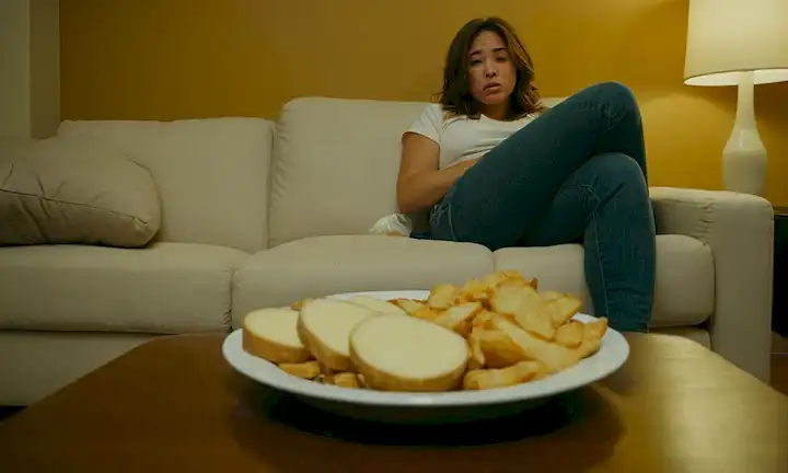 Un plato con una patata parcialmente comida, una escala con pesos desiguales, una persona sentada en un sofá que parece cansado, una basura se llena de aperitivos insalubres, un reloj que muestra una hora tardía, una persona que lucha para salir de la cama, una nevera abastecida con comida basura, una escala de pesaje que se rompe o funciona mal