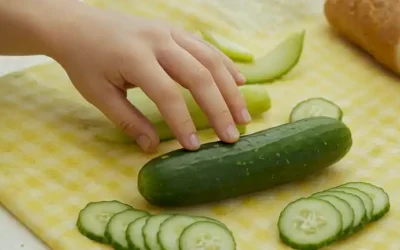Un pepino verde maduro (pepino) sobre un mantel brillante con unas cuantas migajas y papel desgarrado; una mano de niños curiosos que lo alcanza