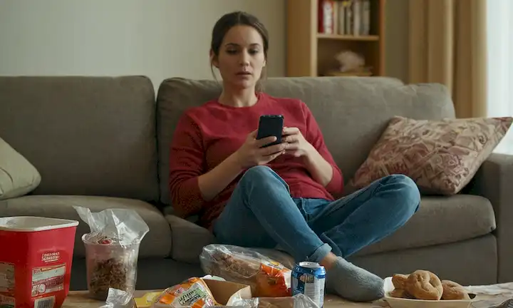 Una persona sentada sola en un sofá con un control remoto de TV, rodeada de envoltorios vacíos de comida y contenedores para llevar, con un fondo borroso de una sala desordenada, usando zapatillas desgastadas, manteniendo un teléfono con un portátil cerrado cerca, con una sensación general de desconexión del mundo