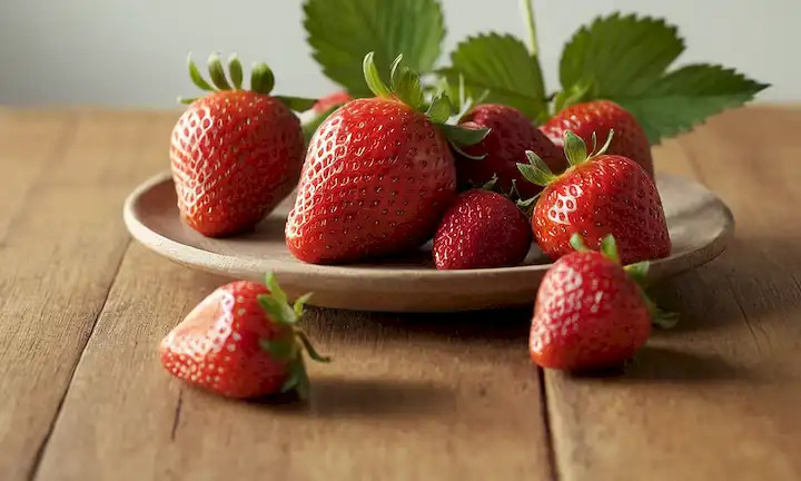 2-3 fresas maduras, algunas hojas y tallos sobre una mesa de madera, luz natural suave, sombras sutiles, fondo ligeramente borroso para enfatizar la fruta