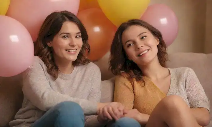 3 mujeres, sonriendo, sentadas juntas en un sofá, sujetando manos o brazos alrededor del otro, rodeadas de flores o globos, iluminación cálida, colores suaves, sin ángulos agudos, entorno natural