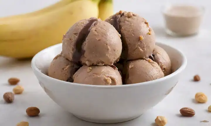 Una cucharada de helado de chocolate de maní de plátano cremoso con un topping natural crujiente en un fondo blanco, con una rodaja de plátano madura, unos cuantos cacahuetes diseminados alrededor, y una gota de salsa de chocolate rica, todo de estilo hermoso contra un entorno limpio y minimalista, capturando los colores vibrantes del postre