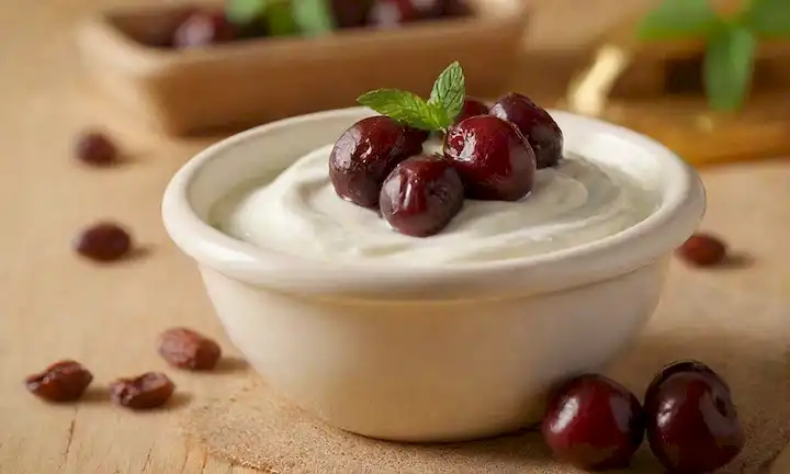 Una base de yogurt cremoso, rematada con cerezas jugosas y fechas caramelizadas, dispersas con hojas de menta frescas, con un fondo dorado cálido, con una cuchara rústica de madera o una cuchara