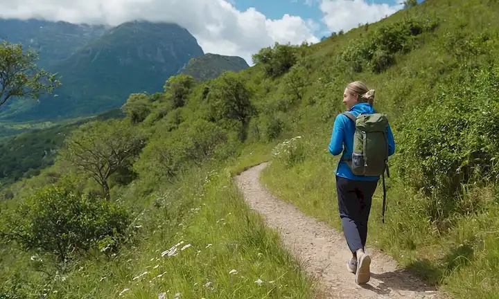 Una persona caminando al aire libre en un sendero pintoresco rodeado de vegetación exuberante, con ropa cómoda y zapatos, con un paso relajado pero deliberado, posiblemente llevando equipaje ligero o botella de agua, con una sonrisa sutil, frente a un cielo azul brillante con nubes blancas suaves, transmitiendo sentido de libertad y alegría