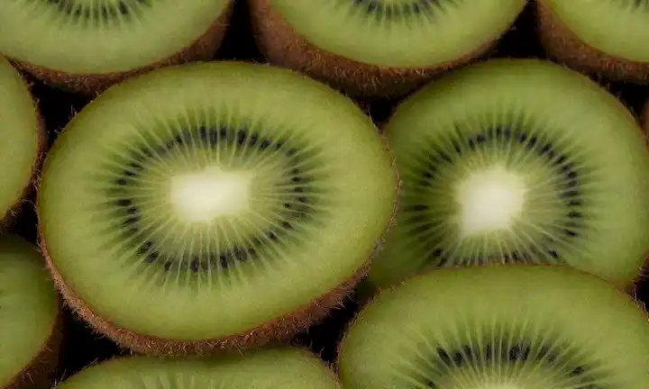 Un kiwifruit vibrante en un fondo blanco, rebanado en la mitad para revelar su carne verde brillante y piel marrón borrosa, con algunas hojas frescas o tallos que la rodean, transportando frescura y bondad natural