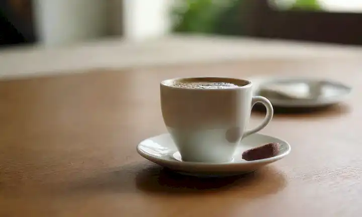 3 kiwis, rebanados y dispuestos en un patrón armónico sobre un mantel de madera; una taza de café caliente al vapor en el fondo, con unas gotas derramadas sobre la mesa; una silueta borrosa débil de una persona sentada en la mesa