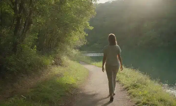 Una persona que camina sobre un sendero escénico en medio de una vegetación exuberante, con un lago o océano sereno en el fondo; una sonrisa sutil y postura relajada que transmite el contenido; la luz del sol caliente que arroja sombras suaves; un sentido de tranquilidad y conexión con la naturaleza