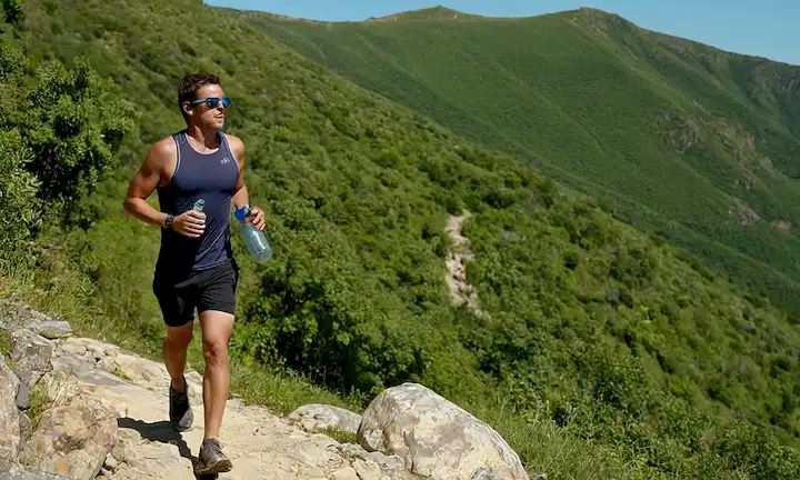5-kilometer trail runner en una ladera soleada con exuberante vegetación y rocas dispersas, con gafas reflectantes de sol, sudor goteando de su frente, mano sosteniendo una botella de agua, piernas en movimiento