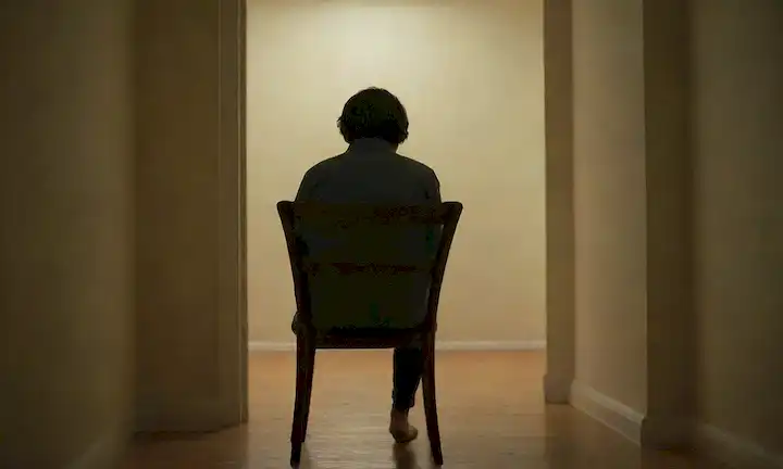 Una persona sentada sola en una silla, rodeada de habitaciones vacías, con una luz débil en el fondo, mostrando un sentido de aislamiento, soledad e introspección silenciosa