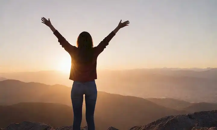 Una persona de pie en un pico de montaña con brazos extendidos a los lados, representando la libertad y el empoderamiento; un atardecer o amanecer en el fondo simbolizando nuevos comienzos; una brisa suave frotando su pelo, significando calma y serenidad; y una sonrisa sutil en su cara, indicando felicidad y cumplimiento