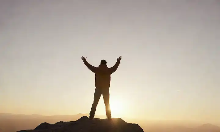 Una persona de pie en un pico de montaña con brazos levantados en el triunfo, amanecer o puesta del sol, luz dorada cálida, niebla sutil, figura inspiradora en movimiento, pose dinámica, transmisión de libertad y empoderamiento