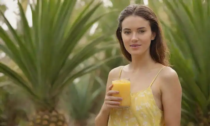 Un ambiente tropical brillante y colorido, un refrescante cristal de zumo lleno de rodajas de piña, una joven que se adapta a una piña fresca, una playa sutil o fondo de jardín con palmeras y flores, la luz del sol caliente filtrando a través de hojas, invitando y sana vibraciones