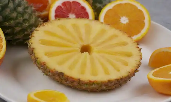 Una rodaja de piña vibrante, fresca y verde, colocada sobre una placa blanca o fondo, rodeada de unas cuantas rodajas de fruta naranja, amarilla o roja para añadir atractivo visual, con luz natural iluminando la escena