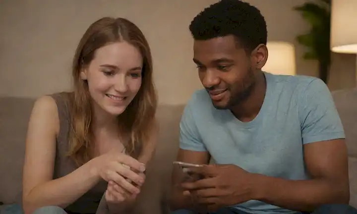 Una joven pareja adulta sentada en un sofá con portátiles y teléfonos inteligentes, rodeada de iluminación suave; Están mirando cada uno de los ojos, con una sonrisa sutil y un lenguaje corporal relajado; Sus dedos están entrelazados, simbolizando la conexión y la intimidad