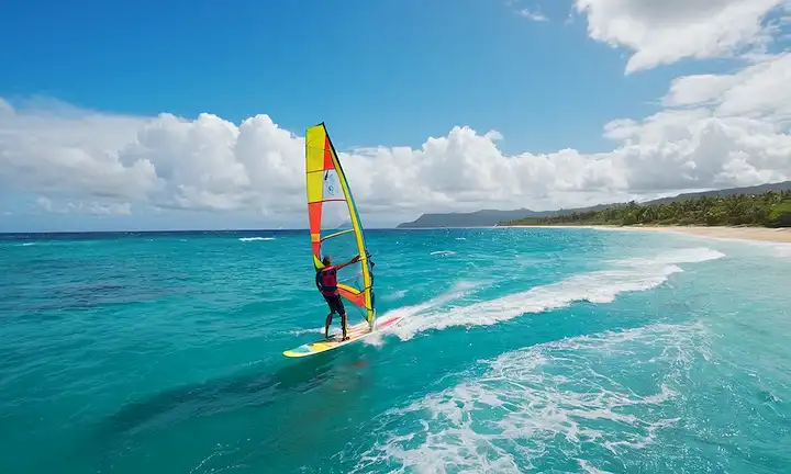 Aguas oceánicas azules claras, un windsurf en movimiento con una sonrisa, velero con diseño colorido, brisa marina soplando, olas chocando, paisajes tropicales, sol filtrando por nubes