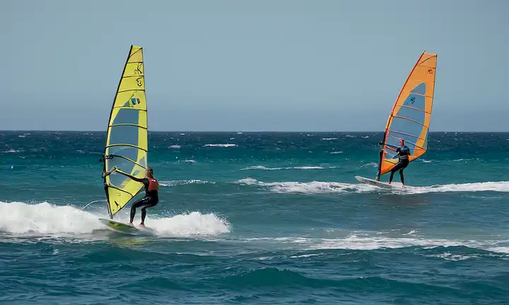 3 windsurfers de pie sobre tablas en aguas poco profundas, otra montando una ola, una voladora con vela extendida, otra balanceando a bordo en mar choppy, una haciendo salto de rampa, y un tiro separado de vela en foco
