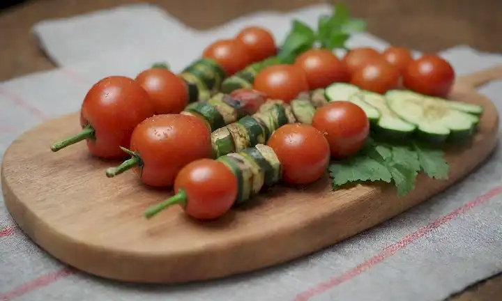 Un arreglo colorido de brochetes en una placa o tabla de madera, con tomates frescos y rebanadas de calabacín, posiblemente algunas hojas de menta, una gota de aceite de oliva, y una rocia de perejil, contra un fondo cálido y natural como un mantel rústico o una superficie de madera