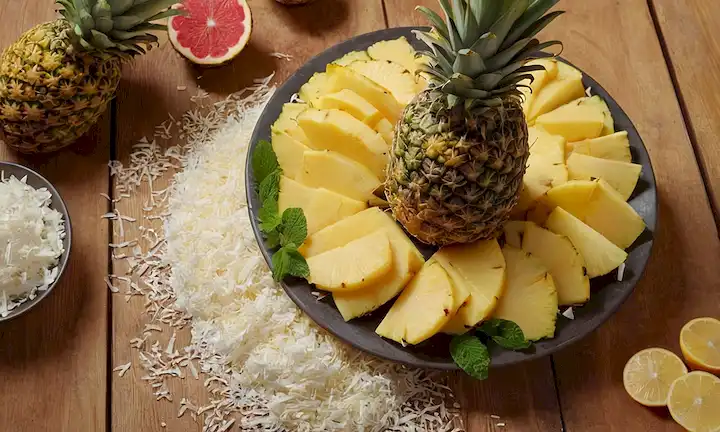 Disposición 3D de frutas coloridas, chirimoya como pieza central, rodeada de anillos jugosos de piña y copos de coco triturados, adornados con hojas de menta frescas en una mesa de madera rústica, luz de oro caliente brillando desde arriba, enfoque suave en primer plano para enfatizar la textura y los colores