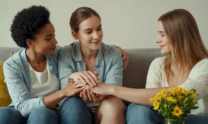 3 personas de diferentes orígenes sentados cómodamente en un sofá, con sus manos tocando o sosteniendo a los demás, rodeados de flores y plantas, transmitiendo confianza y afecto