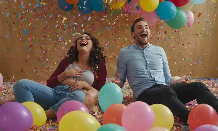 2 personas riéndose, caras contorsionadas, manos en campanas, una con ojos cerrados, otra con boca abierta, tanto en un entorno brillante y colorido, rodeado de globos, confeti y cuerda tonta, con un toque de caos y juguetón