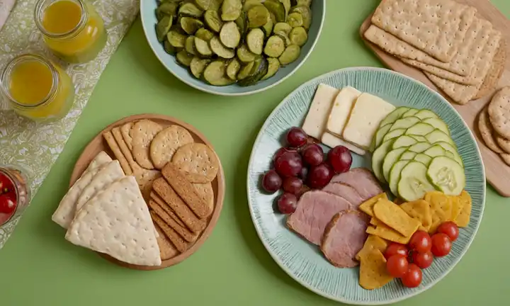 Un plato colorido con una variedad de carnes, quesos y verduras, frutas frescas, galletas de grano entero, un frasco de vidrio lleno de pepinillos, y un fondo verde con un patrón sutil de hojas