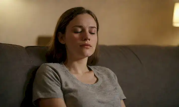 Una persona sentada en un sofá, brazos relajados, ojos cerrados, rodeada de sombras borrosas, con una luz débil en el fondo, mostrando vulnerabilidad, apertura e introspección
