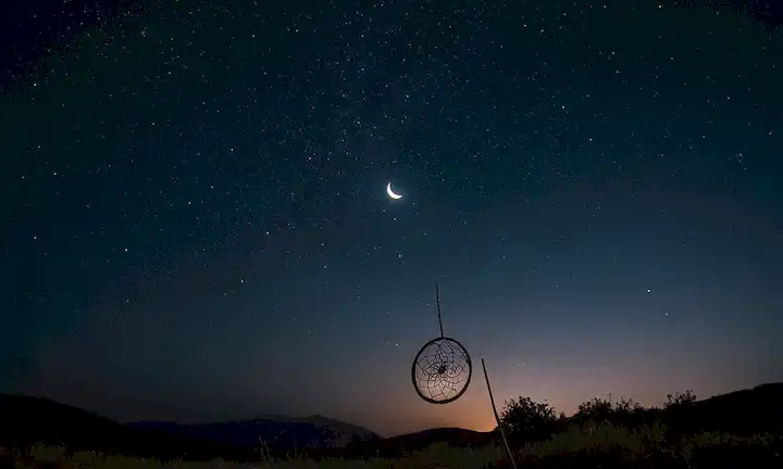 Un cielo oscuro con estrellas dispersas, una luna crescente y un cazador de sueños en el centro, rodeado de nubes brillantes, plumas delicadas y un brillo sutil