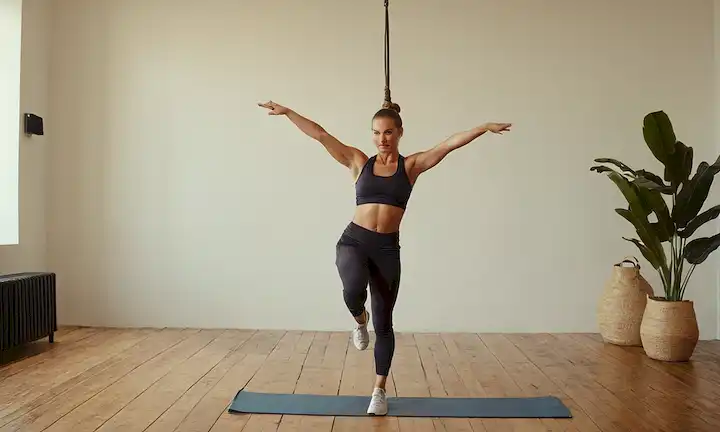 Una mujer en una pose dinámica, de pie en un suelo de madera, usando ropa deportiva, con su pelo atado hacia atrás, mostrando control y confianza; brazos extendidos para demostrar varias posiciones dominantes, con cada mano agarrando una barra o cuerda imaginaria; sudor sutil brilla en su piel, indicando un entrenamiento desafiante