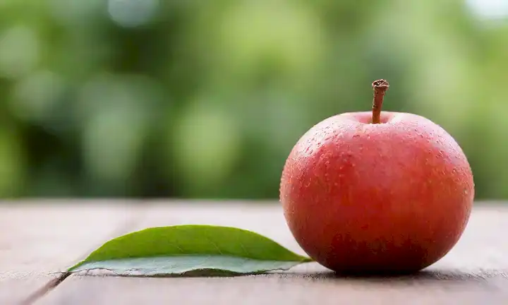 fruta caqi madura sobre una mesa de madera, iluminación natural, fondo borroso, textura fresca y jugosa, ligeras imperfecciones en la piel, hojas verdes vibrantes, pocos tallos dispersos alrededor de la fruta, colores cálidos